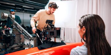 A woman sits with her back to the viewer and is filmed by a man with a camera.