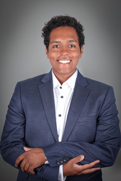 A coloured man in a suit stands in front of a grey background and looks smilingly into the camera, his arms crossed.