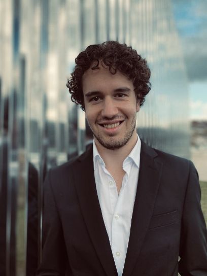 Frontales Portait von Mann mit Locken im Anzug. Leichter Bart, lacht in die Kamera. Im Hintergrund ein reflektierendes Gebäude.