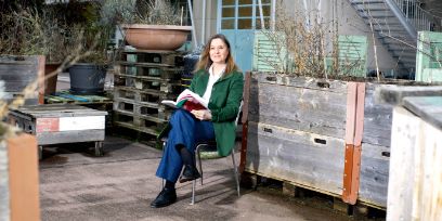 Eine Frau sitzt draussen vor einem Gebäude auf einem Stuhl und hat ein Buch in der Hand. Sie trägt eine grüne Jacke, blaue Jeans und blickt in die Kamera.