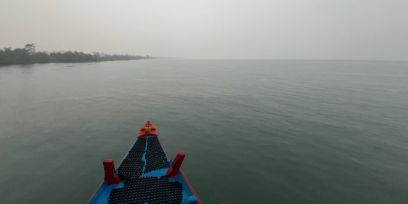 Aus der Sicht einer auf dem Schiff sitzenden Person: Spitze des Schiffes auf gräulichem Wasser. Am Horizont links ist etwas Land zu sehen. Auch der Himmel ist eher grau.